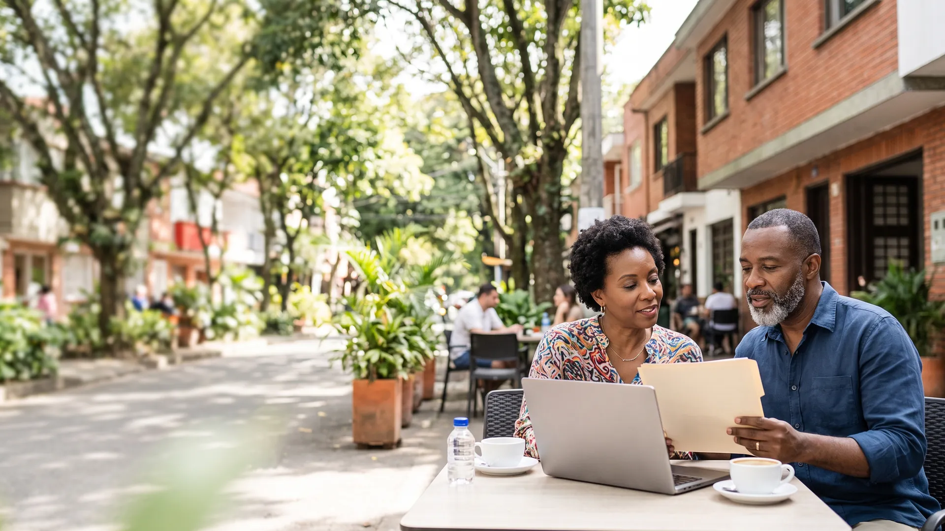 A couple reviews a folder of paperwork and a laptop at a sunny sidewalk café in Medellín